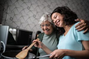 An older adult receiving home care services in Chicago smiles and hugs her caregiver as the caregiver stirs a meal over the stove. 