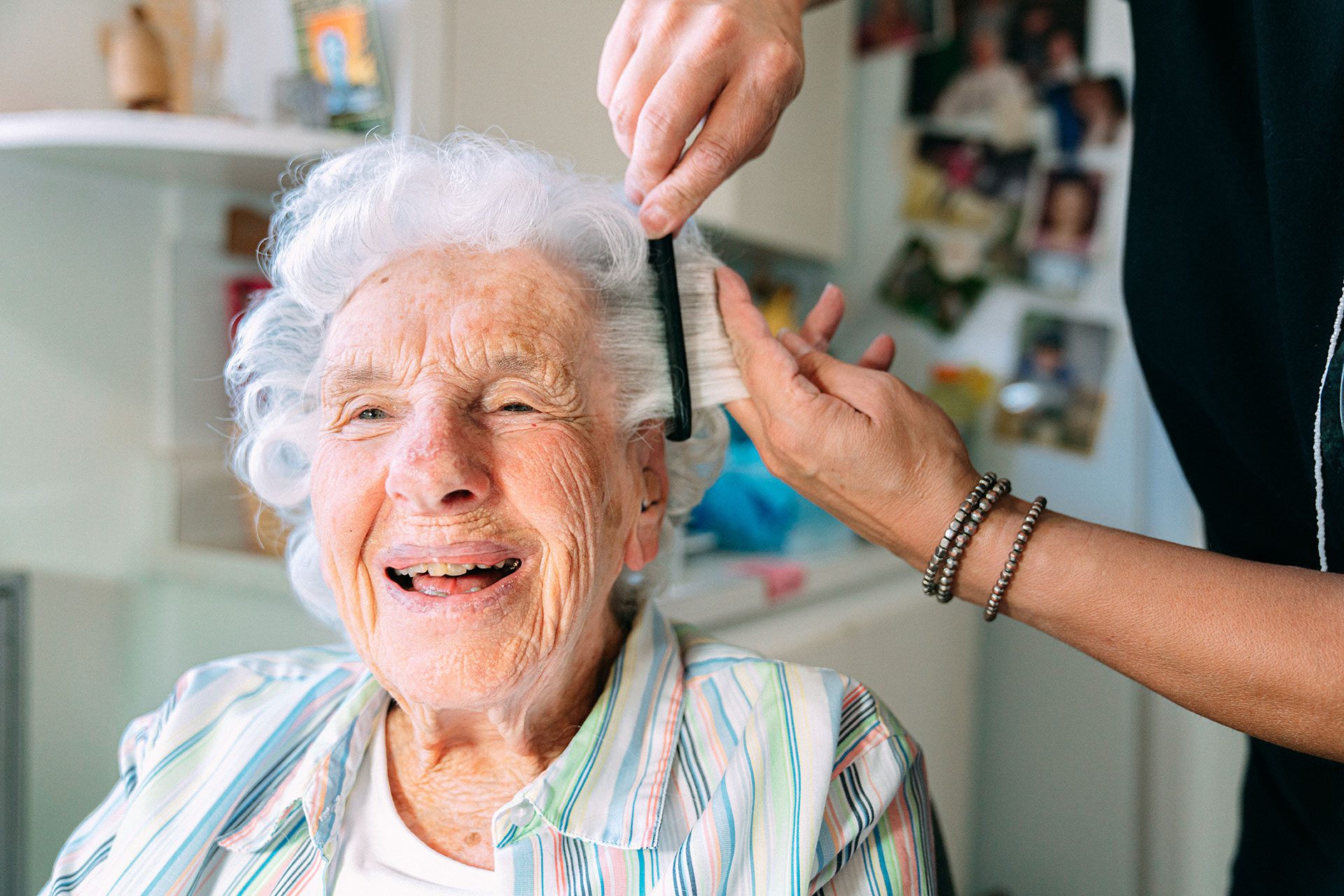 An older woman receiving personal care services at home smiles as her caregiver combs her hair.